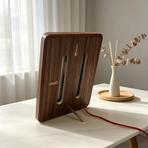 May include: A rectangular dark wood frame with rounded corners, featuring two vertical cutouts and a gold-toned metal kickstand. A thin red power cord extends from the bottom edge of the frame, which is positioned at an angle on a surface.