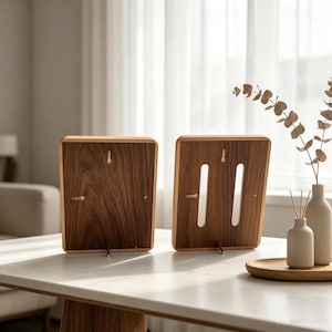 May include: Two rectangular wooden picture frames shown from the back, featuring dark brown wood grain, metal swivel clips for securing contents, and a small metal stand attached to the bottom edge of each frame for upright display on a flat surface.