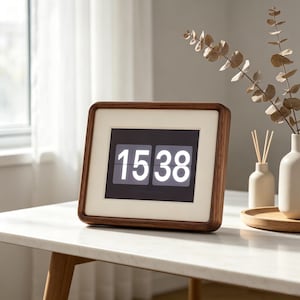 May include: A rectangular digital flip clock with a dark wood frame and a cream-colored mat border. The black display screen shows the white numerical time 15:38. The clock has rounded corners and is positioned on a white surface.