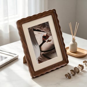 Può includere: Una cornice portafoto rettangolare con finitura in venature del legno marrone scuro e un bordo ondulato smerlato. La cornice presenta un passe-partout color crema e il testo "Muir Wedding" inciso sul bordo inferiore, mostrando una foto in bianco e nero.