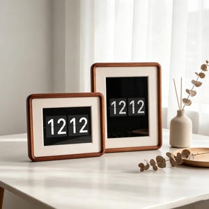 Könnte beinhalten: Zwei rechteckige digitale Klappzahlenuhren mit dunklen Holzrahmen und weißem Passepartout. Die Uhren haben schwarze Displays, die die Uhrzeit 12:12 in weißen Ziffern anzeigen. Eine Uhr ist horizontal, die andere vertikal ausgerichtet.