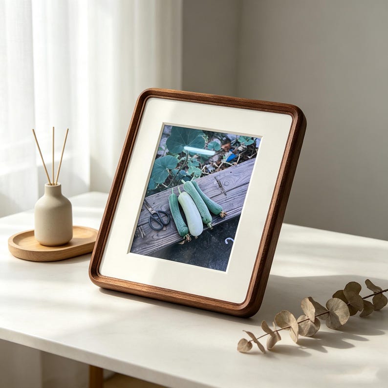 May include: A square wooden picture frame with a cream-colored mat board containing a photograph of three green zucchini resting on a wooden surface next to a pair of black scissors. The frame has rounded corners and a dark brown wood grain finish.