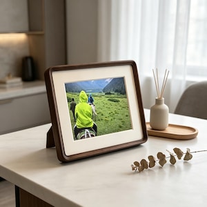 May include: A rectangular dark wood picture frame with a cream-colored mat, displaying a photograph of individuals riding horses through a green mountain landscape. The frame features rounded corners and a stand on the back for tabletop display.