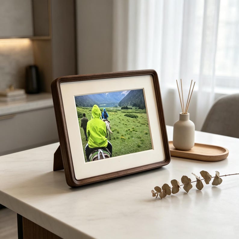 May include: A rectangular dark wood picture frame with a cream-colored mat, displaying a photograph of individuals riding horses through a green mountain landscape. The frame features rounded corners and a stand on the back for tabletop display.