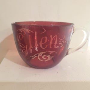 May include: A deep red glass teacup featuring the name Ellen etched in a decorative script font on the front surface. The cup has a smooth, rounded bowl shape and a clear, translucent glass handle attached to the right side of the vessel.