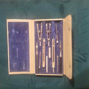 Puede incluir: Un juego de instrumentos de dibujo técnico vintage en un estuche plegable de color crema con interior de terciopelo azul. El set incluye varios compases, divisores y herramientas de dibujo de metal plateado en compartimentos.
