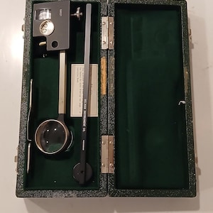 Puede incluir: Un planímetro de Gelman Instrument Co. en un estuche verde oscuro con bisagras y forro de terciopelo. La herramienta cuenta con un cuerpo de metal gris, una lente de aumento circular y un brazo negro largo con el texto 56021 y Made in Germany.