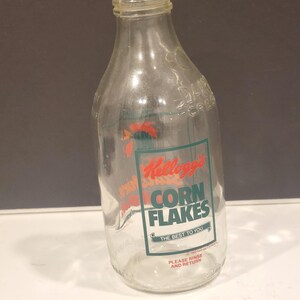 Könnte beinhalten: Eine durchsichtige Glasflasche mit einer Kellogg's Corn Flakes Grafik in Rot und Grün. Der Text lautet Kellogg's CORN FLAKES, THE BEST TO YOU und PLEASE RINSE AND RETURN. Die Flasche hat einen schmalen Hals und einen breiten Boden.