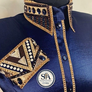May include: A navy blue long-sleeve shirt featuring a high collar and cuffs heavily embellished with gold, dark blue, and clear crystals in geometric patterns. The button placket is lined with gold trim and dark blue buttons. A logo reads SA INTERNATIONAL.