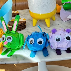 Op de afbeelding: Een verzameling van zes kleurrijke papieren figuren met het label CRUMPIES, voorzien van wiebelogen en diverse vormen, waaronder een roze varken, geel wezen, groen konijn, blauw figuur, paars personage en een wit wezen met pompons.