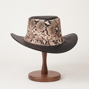 May include: A cowboy hat featuring a black crown top and a wide black brim. The side of the crown displays a tan and brown snakeskin pattern with a band accented by small metallic eyelets encircling the base of the crown above the brim.