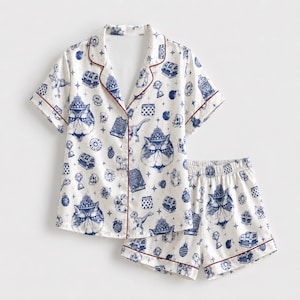 May include: A white pajama set with short sleeves and shorts, featuring a blue repeating pattern of cats wearing crowns, treasure chests, skulls, dinosaurs, and trophies. The shirt has a button-down front and thin red piping along the edges.