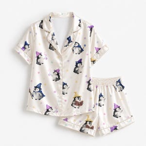 May include: A cream-colored short-sleeve pajama set featuring a button-down shirt and matching shorts. The fabric is decorated with a repeating pattern of cartoon penguins wearing wizard hats and robes, interspersed with small colorful stars.