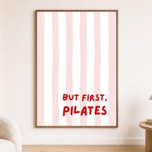 May include: A rectangular wall art print in a wooden frame featuring vertical light pink and white stripes. The text "BUT FIRST, PILATES" is printed in a bold, red, hand-drawn style font in the lower centre of the vertical striped background.