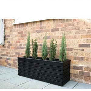 May include: A rectangular black wooden planter box featuring horizontal slats, containing five slender, upright green coniferous plants. The planter is positioned on a light grey tiled surface against a brown brick wall exterior.