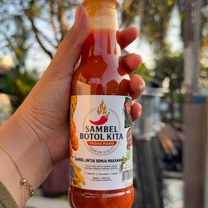 Peut inclure: Une bouteille en plastique transparent remplie de sauce chili orange-rouge, avec un bouchon blanc et une étiquette indiquant SAMBEL BOTOL KITA PEDAS MANIS PREMIUM QUALITY, avec un volume net de 250 ml imprimé sur le bas de l'étiquette.