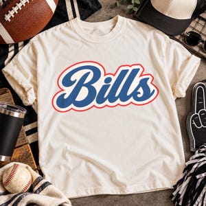 May include: A cream-coloured short-sleeve t-shirt featuring the word Bills in a bold blue script font with a red outline centred on the chest. The shirt is made of a soft fabric and has a classic crew neckline and standard fit.