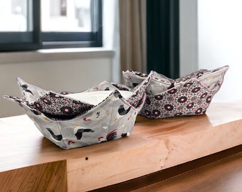 May include: Two fabric bowl cozies on a wooden table. One cozy is gray with a rooster pattern and the other is black and white with a floral pattern.
