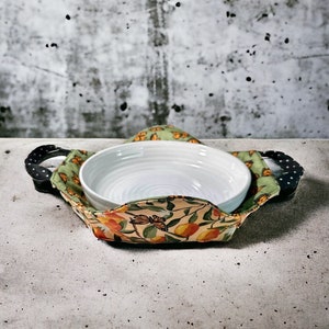 May include: A white ceramic bowl with a green and orange floral print fabric bowl holder. The holder has black handles and a butterfly design.