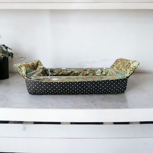 Puede incluir: Un protector de horno de tela negro con lunares blancos y estampado de mariposas verdes y naranjas. El protector está diseñado para proteger los moldes para hornear de arañazos y manchas.