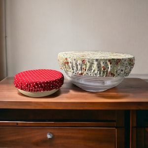 Op de afbeelding: Twee stoffen kom deksels op een houten tafel. De grotere deksel is groen met een bloemenpatroon en de kleinere deksel is rood met witte stippen.