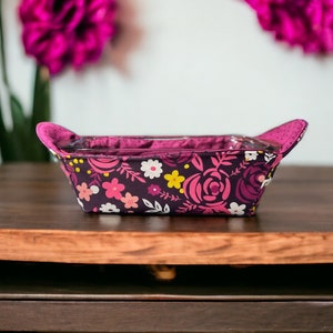 May include: A purple and pink floral patterned fabric baking dish liner with two handles. The liner is sitting on a wooden table.