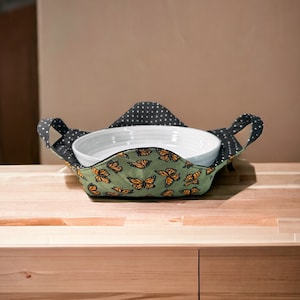 May include: A green fabric dish holder with a black and white polka dot lining and butterfly print. The holder has two handles and is holding a white ceramic bowl.