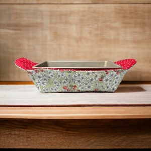 May include: A green and white floral patterned fabric baking dish holder with red polka dot handles. The holder is holding a silver baking dish.