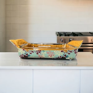 May include: A glass baking dish with a floral print fabric cover. The cover has a yellow trim and is designed to protect hands from heat.