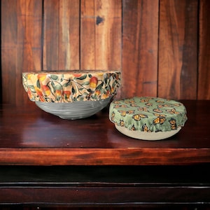 May include: Two reusable food covers, one with a floral pattern and the other with a butterfly pattern, on a wooden surface.
