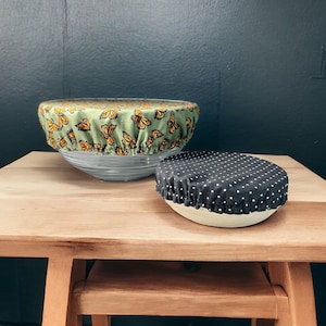 Op de afbeelding: Twee stoffen kom deksels, een met een groene achtergrond en een vlinderpatroon, en de andere met een zwarte achtergrond en witte stippen. De deksels liggen op een houten tafel.
