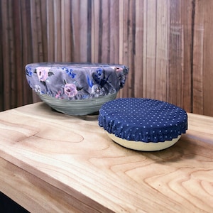 Op de afbeelding: Twee herbruikbare stoffen kom deksels. Eén is grijs met een bloemenpatroon en de andere is donkerblauw met witte stippen.