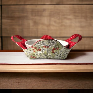 Puede incluir: Un calientaplatos de tela rojo y blanco con un patrón floral y lunares rojos. El calientaplatos tiene dos asas y sostiene un plato blanco.