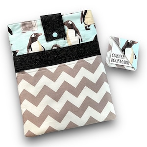 Puede incluir: Una funda para libros con un patrón de pingüinos sobre un fondo azul claro. La funda tiene una cremallera negra y un patrón de chevron negro en la parte inferior. Viene con un marcapáginas de esquina de pingüino a juego que dice "Corner Bookmark".