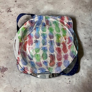 Puede incluir: Un plato para tartas de vidrio con una cubierta de tela con estampado de piña en tonos rojos, azules, verdes y amarillos. La cubierta de tela tiene un ribete de lunares azules y blancos.
