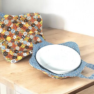 Puede incluir: Dos soportes para platos de tela. Uno es azul con lunares blancos y el otro es un mosaico de amarillo, naranja y marrón. Ambos tienen asas.