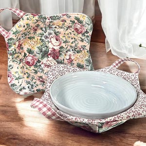 Puede incluir: Dos agarraderas para ollas con estampado floral. Una es una agarradera cuadrada con asa. La otra es una agarradera redonda con un tazón dentro.