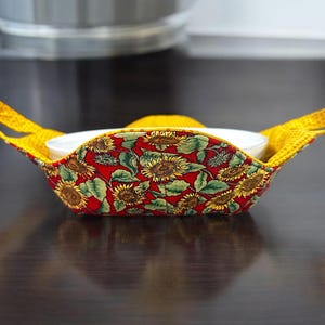 Puede incluir: Un bol de tela amarillo y rojo con estampado de girasol y asas. El bol contiene un bol de cerámica blanco.