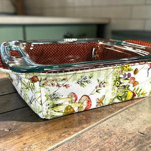 May include: A rectangular glass baking dish with a colorful fabric cozy. The cozy features a white background with a floral and mushroom pattern. The handles are covered in a red fabric with white polka dots. The dish sits on a wooden surface.