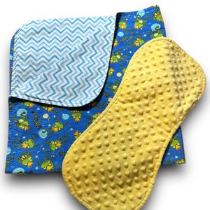 May include: A blue and white chevron patterned baby blanket with a yellow burp cloth featuring a frog print. The frog print includes the words "Little Prince".