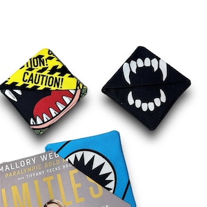 Könnte beinhalten: Drei Stoff-Lesezeichen mit verschiedenen Designs. Ein Lesezeichen ist schwarz mit weißen Zähnen, ein anderes ist schwarz mit einem gelben und schwarzen Warnschild und einem Comic-Monster, und das dritte ist blau mit einer schwarzen Haifischflosse.