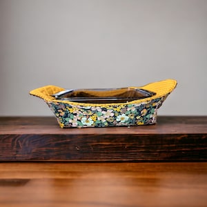 Puede incluir: Un portador de fuente para horno de tela con estampado floral y ribete amarillo. El portador está sobre una superficie de madera marrón oscura.