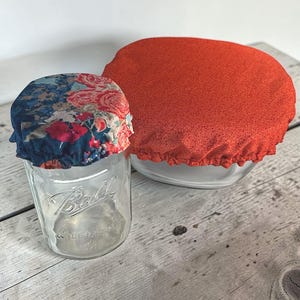 Op de afbeelding: Een glazen pot met een bloemenstoffen deksel naast een glazen kom bedekt met een rode stoffen deksel. Op de pot staat het woord "Ball". De deksels zijn ontworpen om de pot en kom te bedekken.