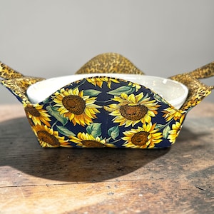 May include: A fabric bowl cosy with a sunflower pattern on a navy blue background. The cosy has leopard print handles and holds a white bowl. The cosy is designed to protect hands from hot bowls.