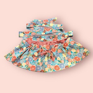May include: A floral print apron with a red and white polka dot trim. The apron has a tie at the waist and is made of a lightweight fabric.