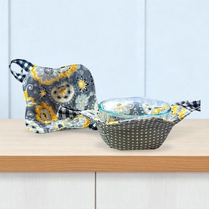 May include: Two fabric bowl cozies with a floral pattern in gray, yellow, and white. One cozy is holding a clear glass bowl. The other cozy is on the left. The cozies have a black and white checkered handle.