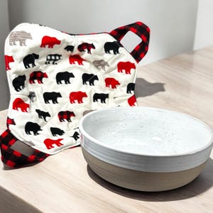 May include: A white ceramic bowl with a speckled glaze sits on a wooden surface next to a red and black quilted hot pad with a pattern of bears.