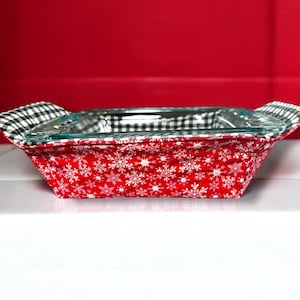 May include: A red and white casserole dish carrier with a snowflake pattern. The carrier has black and white checkered fabric on the handles.