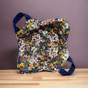 Puede incluir: Una cesta de tela floral azul y blanca con dos asas. La cesta está hecha de una tela suave y acolchada y tiene un patrón floral con flores rosas, amarillas y verdes.