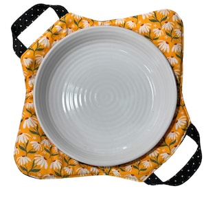 Puede incluir: Un salvamanteles de tela floral amarillo y blanco con asas de lunares negros. El salvamanteles está diseñado para sujetar un plato o un plato.